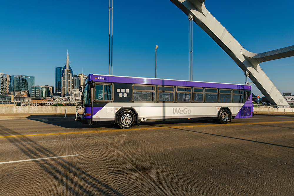 A purple and white WeGo bus crosses a bridge under a clear blue sky. In the background, a city skyline with modern skyscrapers is visible.