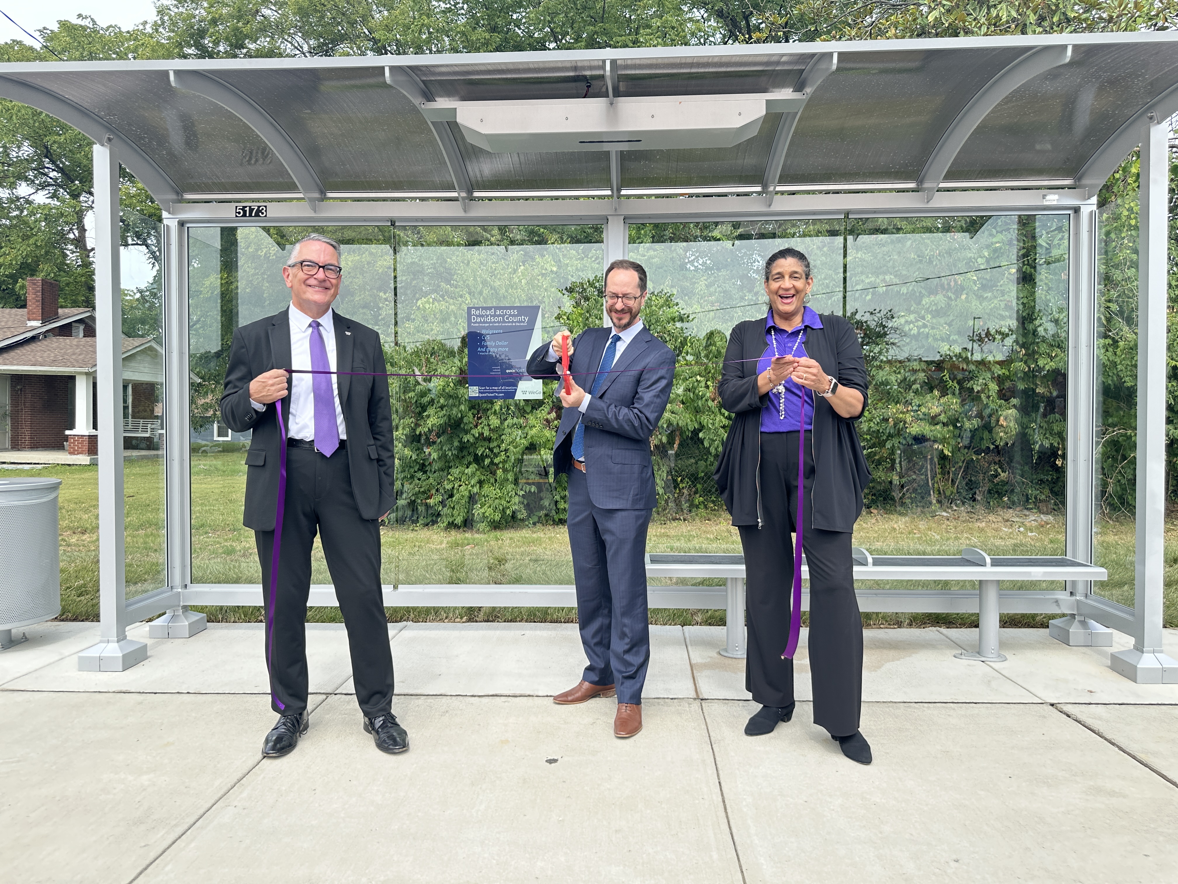 Three people stand smiling at a bus stop ribbon-cutting ceremony
