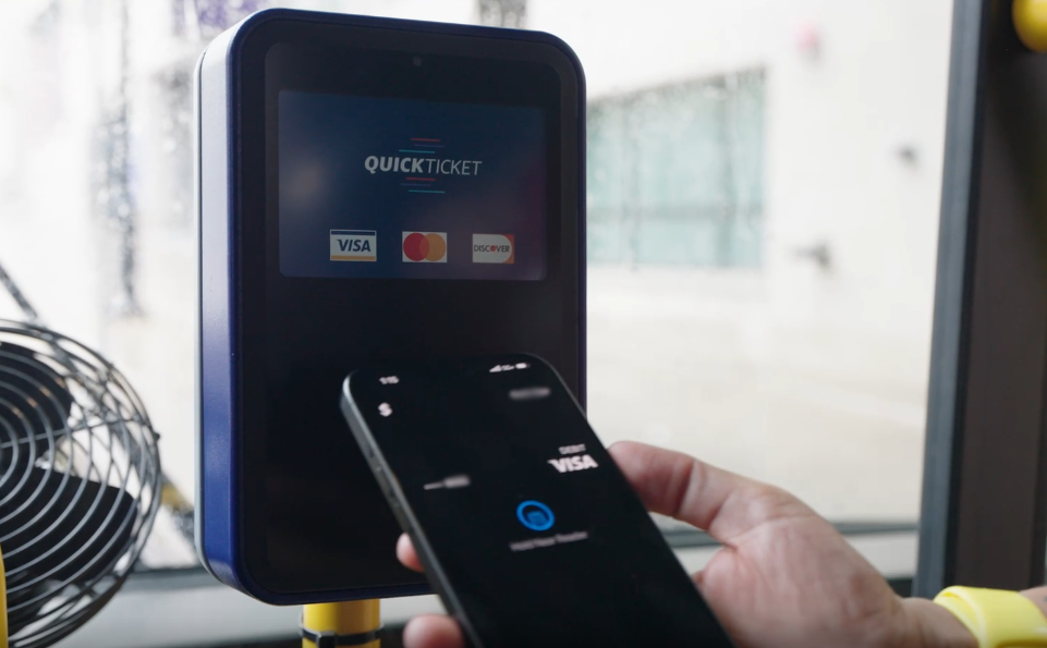 A hand holds a smartphone showing a payment screen near a bus ticket validator with QuickTicket and credit card company logos displayed on the screen