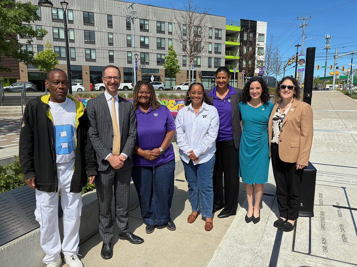A diverse group of seven people smiling outside on a sunny day, with a modern building and colorful mural in the background
