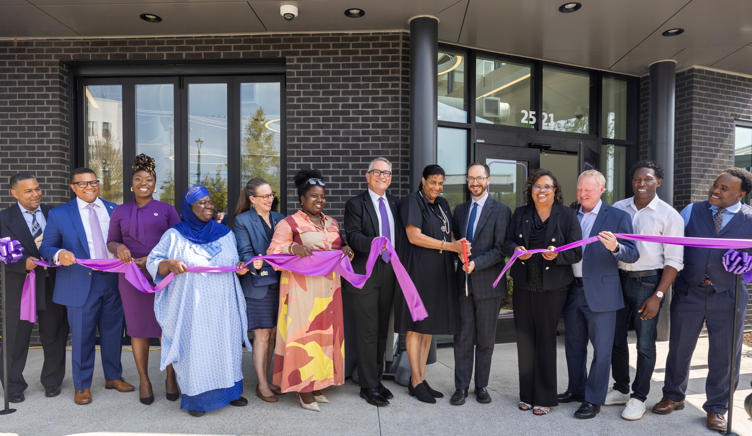 A diverse group of people stands in front of a building, smiling and holding a long purple ribbon. A man in the center holds ceremonial scissors.