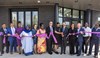 A diverse group of people stands in front of a building, smiling and holding a long purple ribbon. A man in the center holds ceremonial scissors.