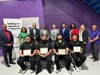 A group of people stand and sit in front of a purple wall. Four seated men hold certificates, with a diverse team behind them, smiling and posing. A banner reads Building the Workforce of Tomorrow.