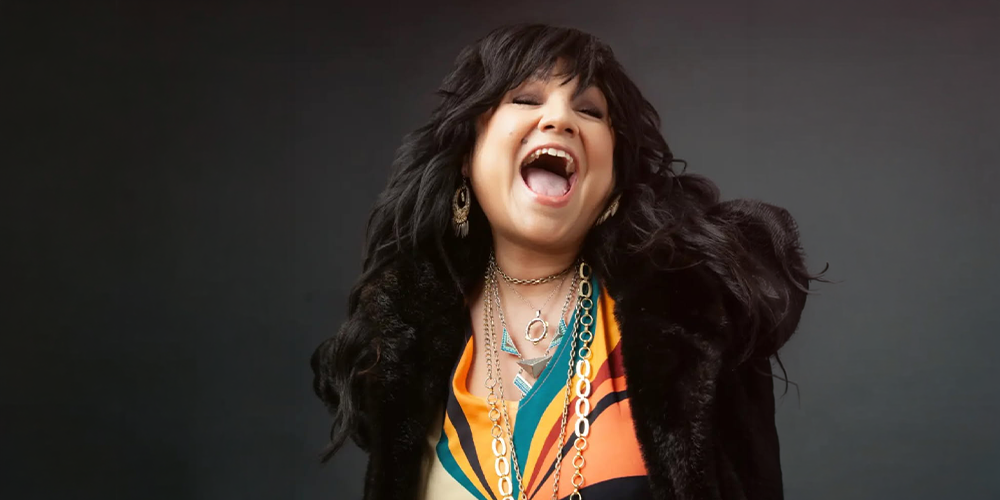 A joyful woman with dark, wavy hair laughs openly, wearing bold, colorful clothing and layered necklaces