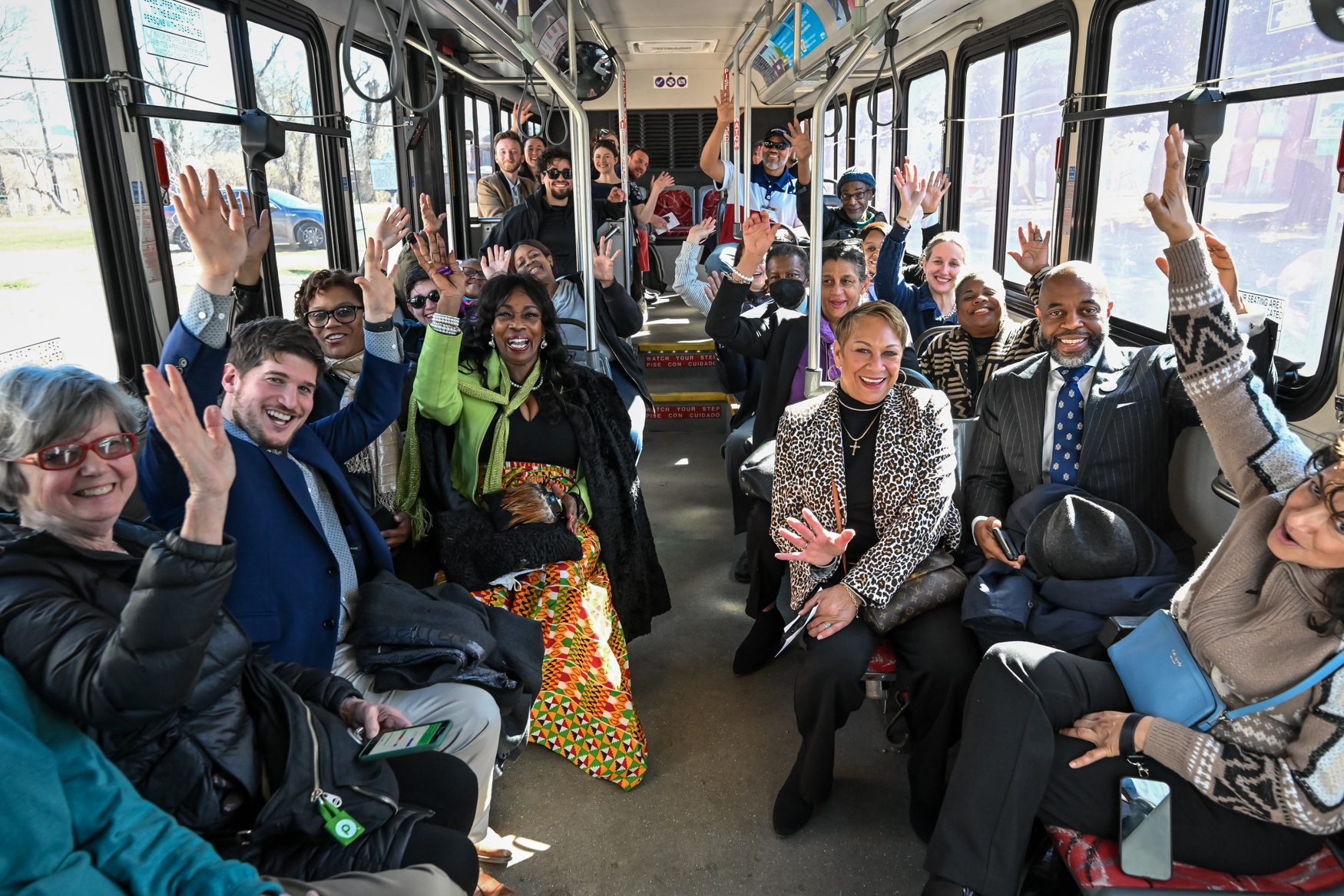 A group of people sit in a bus, smiling and raising their hands in a cheerful, celebratory manner