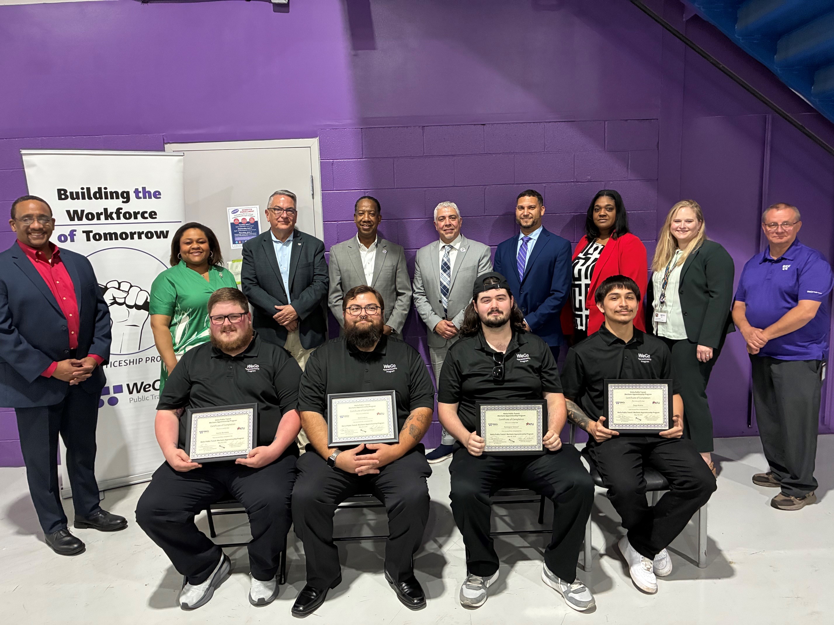 A group of people stand and sit in front of a purple wall. Four seated men hold certificates, with a diverse team behind them, smiling and posing. A banner reads Building the Workforce of Tomorrow.